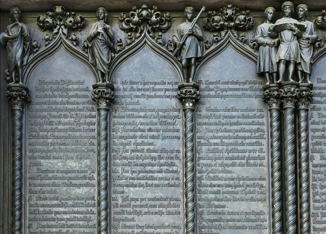 Das Bild zeigt die Thesentür an der restaurierten Schlosskirche in Wittenberg, im Detail sind die 95 Thesen angezeigt und darüber eine Gemälde.