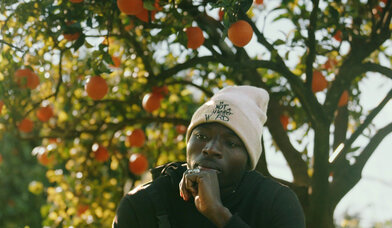 Ein Mann mit weißer Mütze steht vor einem Orangenbaum, an dem Früchte hängen. - Copyright: Marcus Zahn