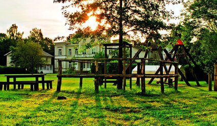 Rasenfläche mit einem eingezäunten Baum, rechts ein Tisch mit Bank, im Hintergrund eine weiße Villa - Copyright:  Emilio_Douglas