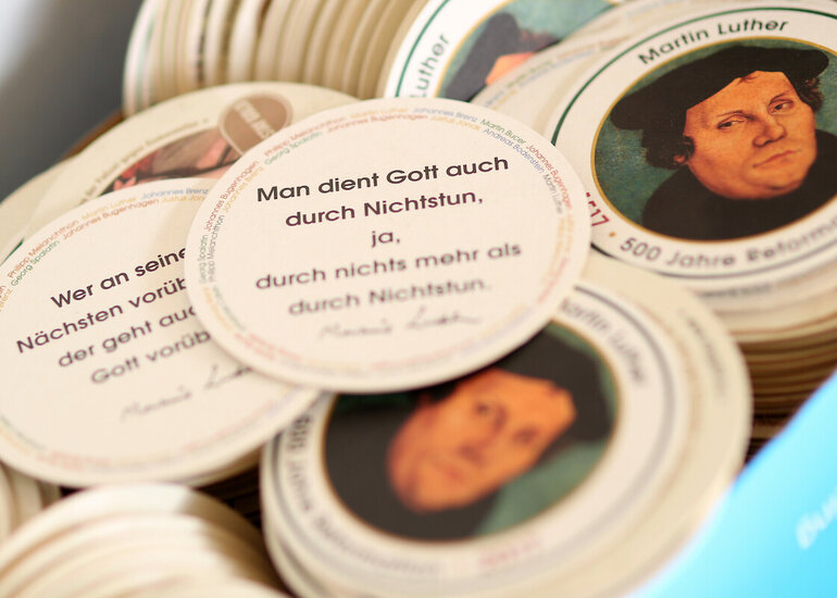 Kiste mit vielen Bierfilzen, einige mit Portrait, andere mit Zitat nach oben sichtbar. Fokus auf eines mit Zitat