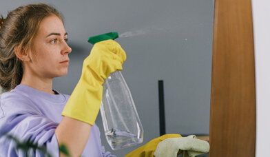 Eine Frau im fliederfarbenen Pullover hält eine Sprühflasche mit Reinigungsmittel in der einen und einen Putzlappen in der anderen Hand. - Copyright: SHVETS/pexels